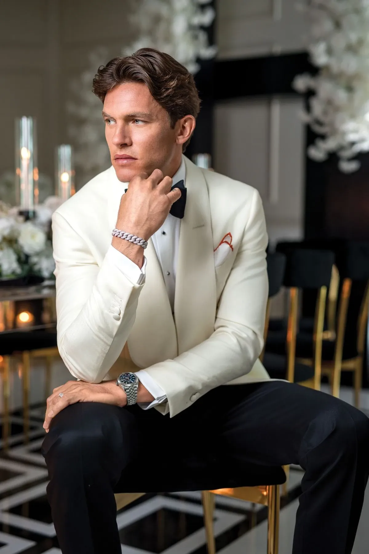A groom is seated in an elegant setting, wearing a stylish ivory tuxedo jacket with a black bow tie. He is adorned with exquisite wedding jewelry from Angelica Jewellers, including a sophisticated watch and a diamond bracelet, highlighting the wedding jewelry category. The backdrop features luxurious wedding decor, including white floral arrangements and softly lit candles, creating a romantic atmosphere ideal for a lavish wedding celebration.