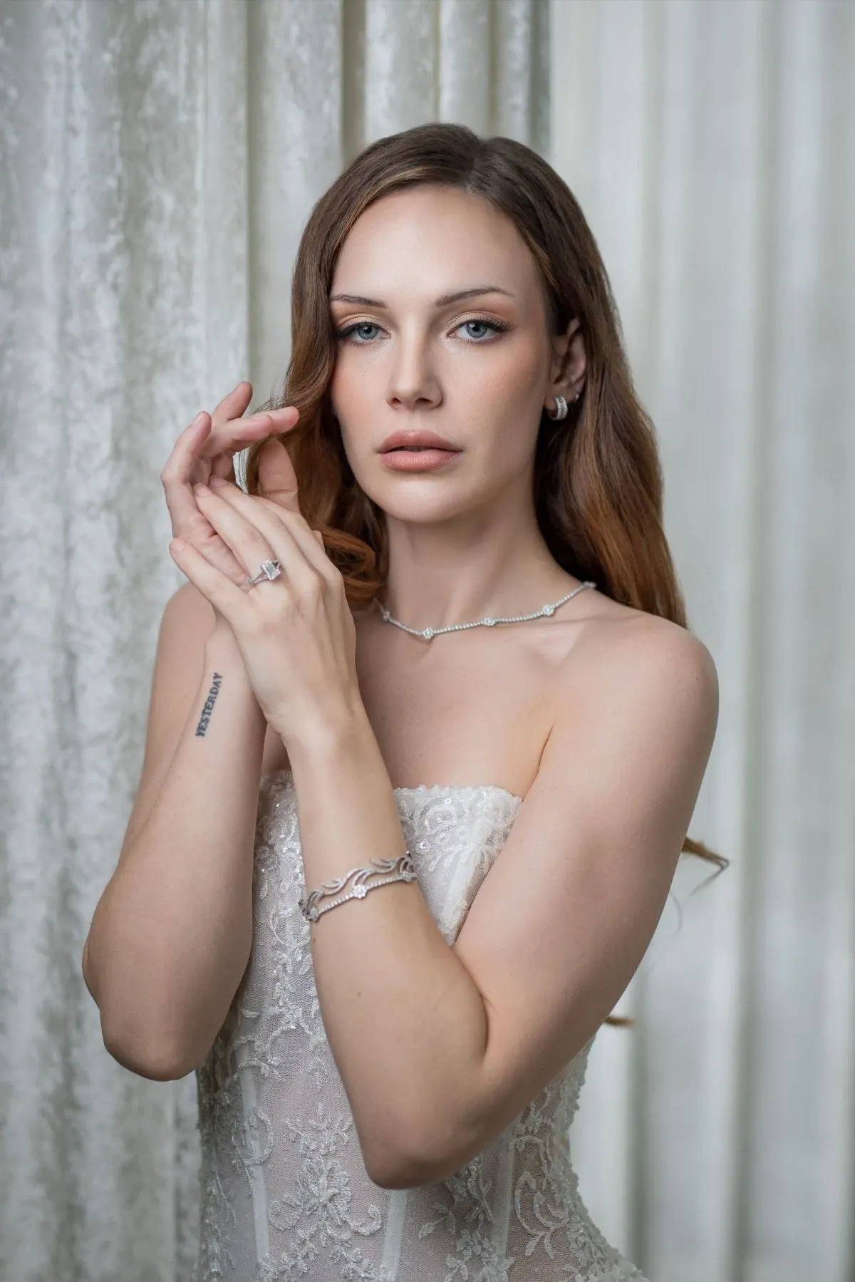 A bride stands gracefully against a neutral, textured backdrop, showcasing elegant bridal jewelry by Angelica Jewellers. She is adorned with a delicate necklace that complements her strapless lace wedding gown. Her subtle yet sophisticated earrings and a matching bracelet enhance the overall bridal look, perfect for the wedding jewelry category. The image captures the essence of elegance and refinement, ideal for brides seeking timeless and beautiful wedding accessories.