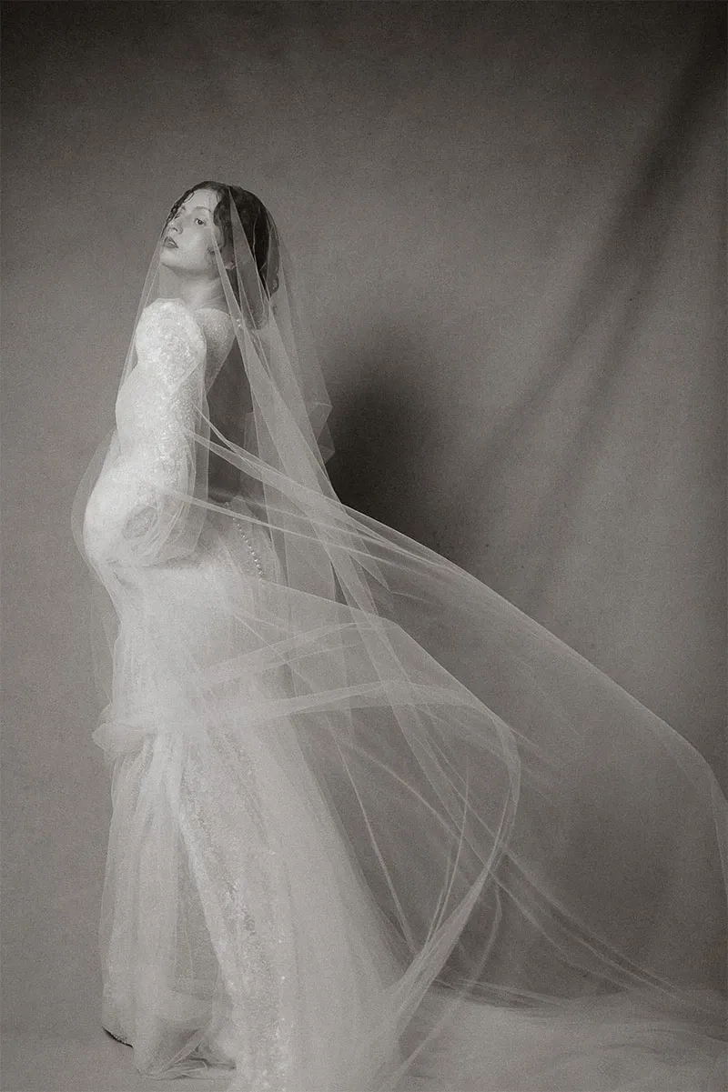 This elegant black and white image, provided by Acrewood under the Wedding Photography category on Mariage Elegant, Canada, showcases a bride dressed in a stunning, form-fitting lace gown. The gown features intricate patterns and is complemented by a long, flowing veil that drapes gracefully over her. The bride stands posed gracefully, adding an ethereal and sophisticated touch to the composition, ideal for a timeless wedding aesthetic.