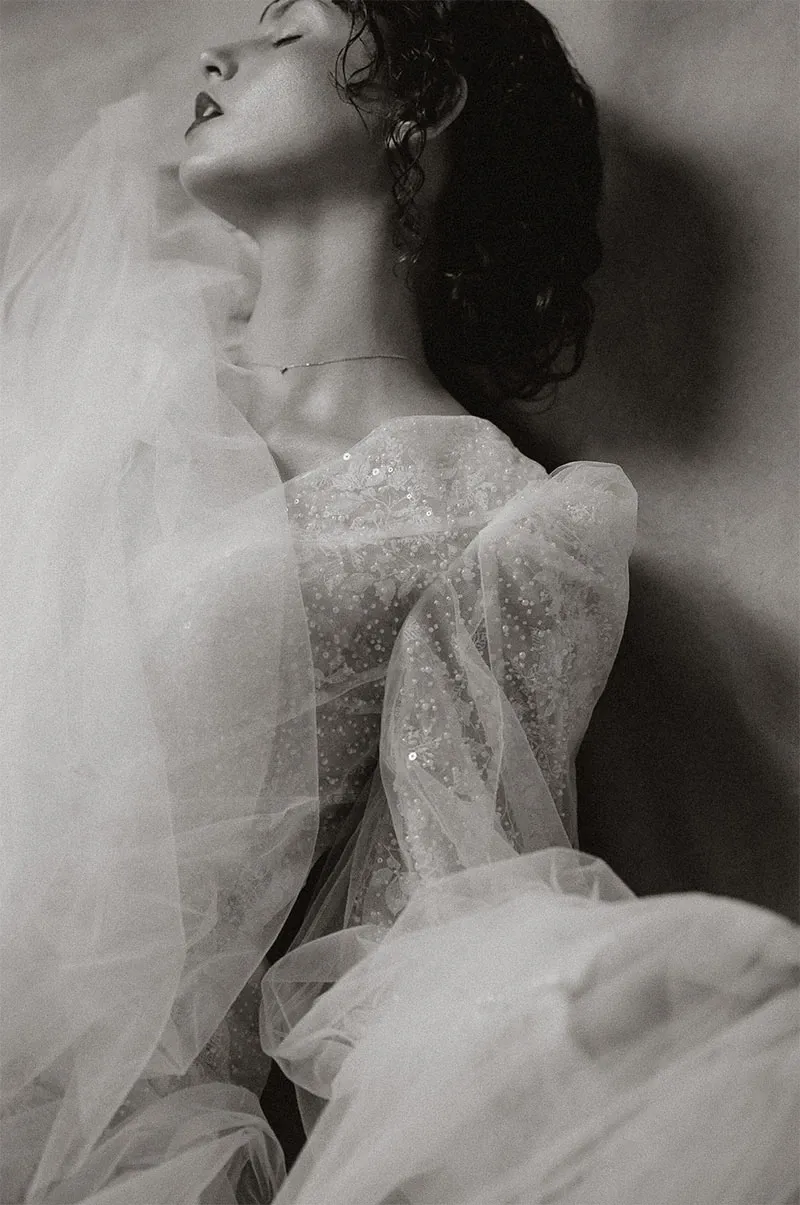 This black and white image, provided by Acrewood, features a bride elegantly captured in the midst of her wedding day, highlighting the vendor's expertise in wedding photography. The bride is wearing a stunning, sequin-embellished gown with sheer, flowing fabric, exuding an ethereal and sophisticated aesthetic. Her hair is styled in soft curls, and she is posed with her head tilted back, showcasing a sense of serene elegance. The photography expertly captures the texture and details of the gown, adding to the timeless and romantic feel.