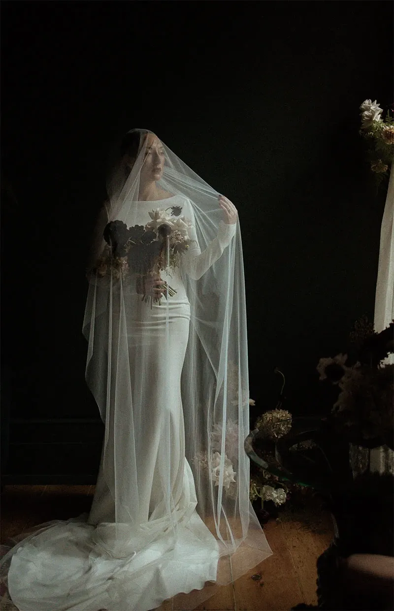 A bride stands elegantly holding a bouquet of flowers, with a sheer veil cascading over her body, set against a dark, moody background. Soft light illuminates her serene expression, highlighting the intricate details of her white gown and the textures of the flowers. This captivating moment is captured by Acrewood, a distinguished vendor in the Wedding Photography category for Mariage Elegant in Canada, showcasing their skill in creating timeless and emotive bridal portraits.