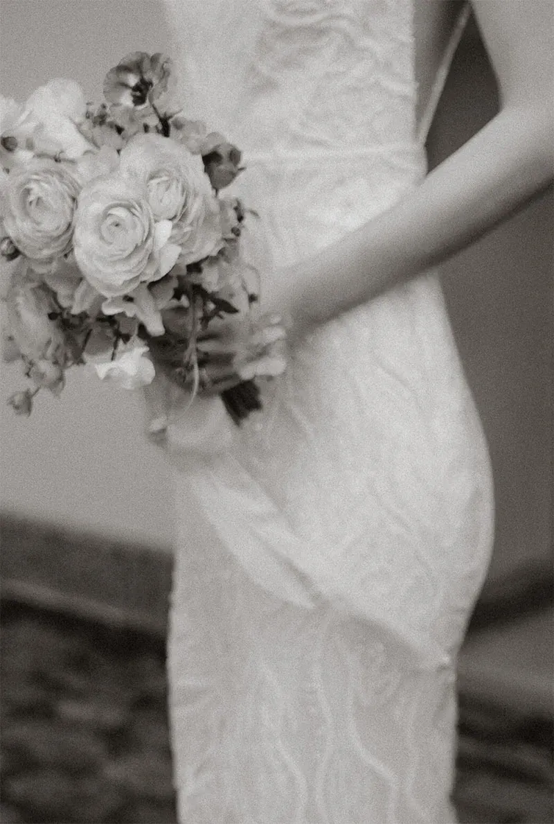 The image shows a bride in a beautifully detailed white lace wedding dress, holding an elegant bouquet of flowers. The bouquet features roses and other delicate blooms, adding a touch of romance and sophistication. This image, captured by Acrewood, showcases their expertise in wedding photography by highlighting the intricate design of the bridal gown and the softness of the floral arrangement. The focus is on the bride's form and the texture of the gown, emphasizing the elegance and grace of the wedding day.
