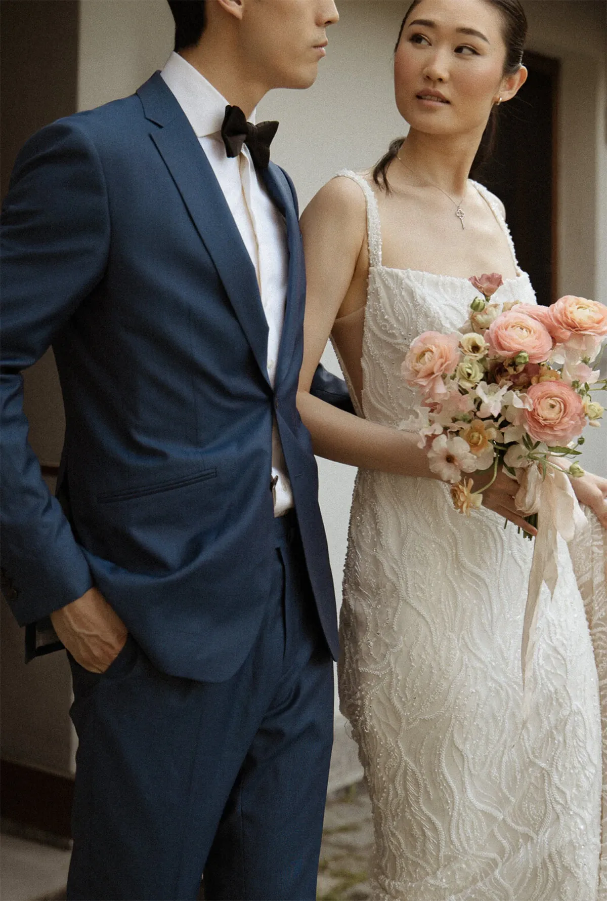 A stylish wedding couple stands in a serene outdoor setting, embodying elegance and romance. The groom is dressed in a tailor-fitted blue suit with a classic black bow tie, exuding sophistication. The bride is adorned in a stunning white beaded gown, holding a delicate bouquet of pink and peach flowers, adding a touch of color and elegance. This image represents the graceful work of Acrewood in the Wedding Photography category, as featured on Mariage Elegant, Canada.