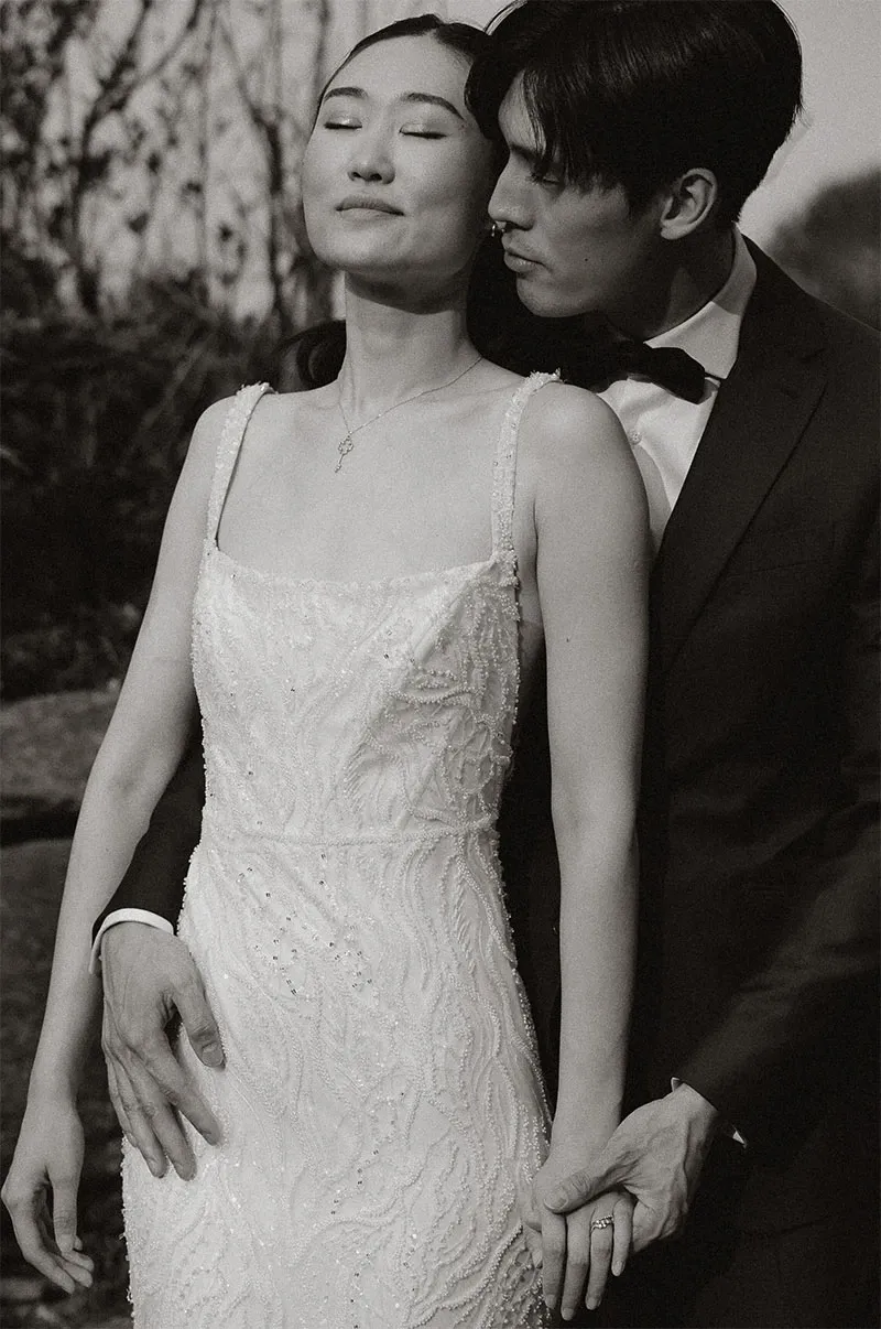 This black and white image features a bride and groom in a serene outdoor setting, captured by Acrewood in the wedding photography category. The bride is wearing a detailed, elegant gown with intricate beadwork and has her eyes closed, exuding a sense of calm and contentment. The groom, dressed in a classic dark suit and bow tie, gently holds her hand and leans in to kiss her forehead, embodying an intimate and tender moment. The background suggests a natural landscape, enhancing the romantic and timeless feel of the photograph, ideal for showcasing Acrewood's skill in capturing authentic wedding moments.