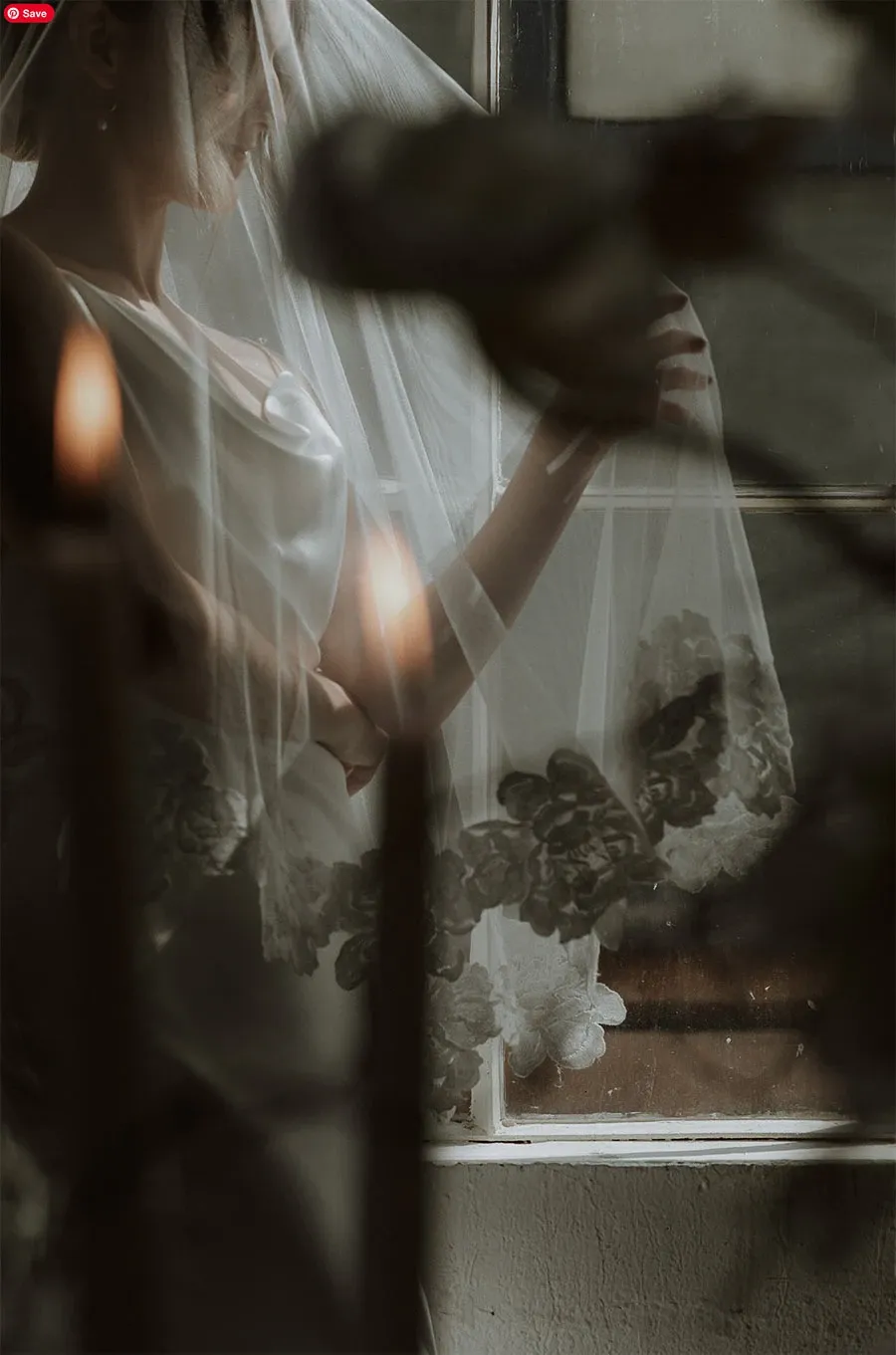 In this ethereal wedding photograph by Acrewood, under the category of Wedding Photography on Mariage Elegant, a bride is captured standing gracefully beside a window. She is partially veiled in delicate lace, its intricate floral patterns softly visible. The ambient lighting casts a warm glow, enhancing the romantic atmosphere as it filters through the window. The blurred foreground elements, which include softly lit candles, add depth and an intimate, dreamy quality to the image, emphasizing the beauty and elegance of the wedding setting.
