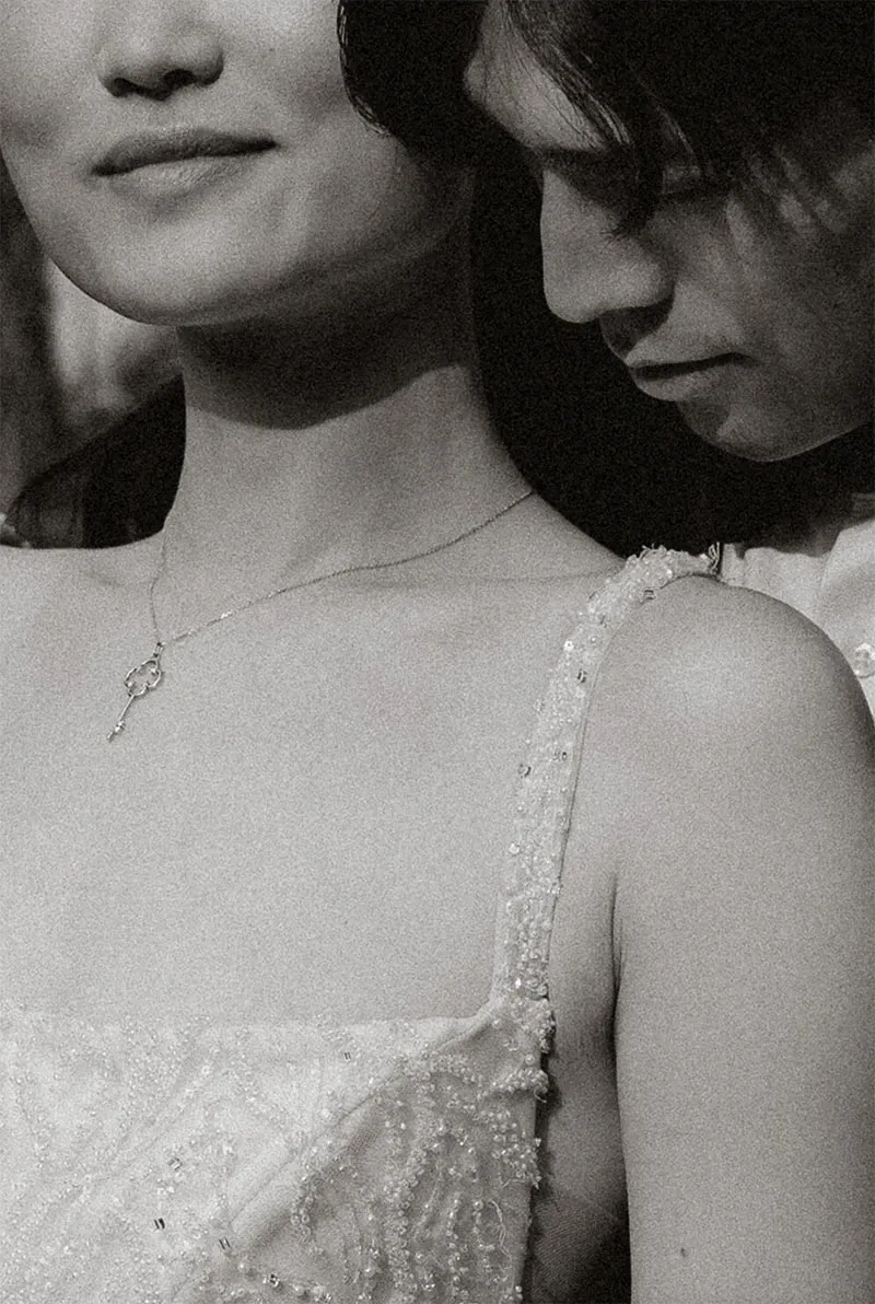 A romantic black and white close-up of a bride and groom, highlighting the elegance of wedding photography by Acrewood, part of the vendor listing at Mariage Elegant, Canada. The bride, wearing an intricately beaded wedding dress, is shown from the shoulders up, with a delicate chain necklace featuring a small pendant. The groom, partially visible, stands closely beside her, resting his head gently against hers. This intimate moment captures the essence of love and grace, ideal for showcasing Acrewood's expertise in capturing timeless wedding memories.
