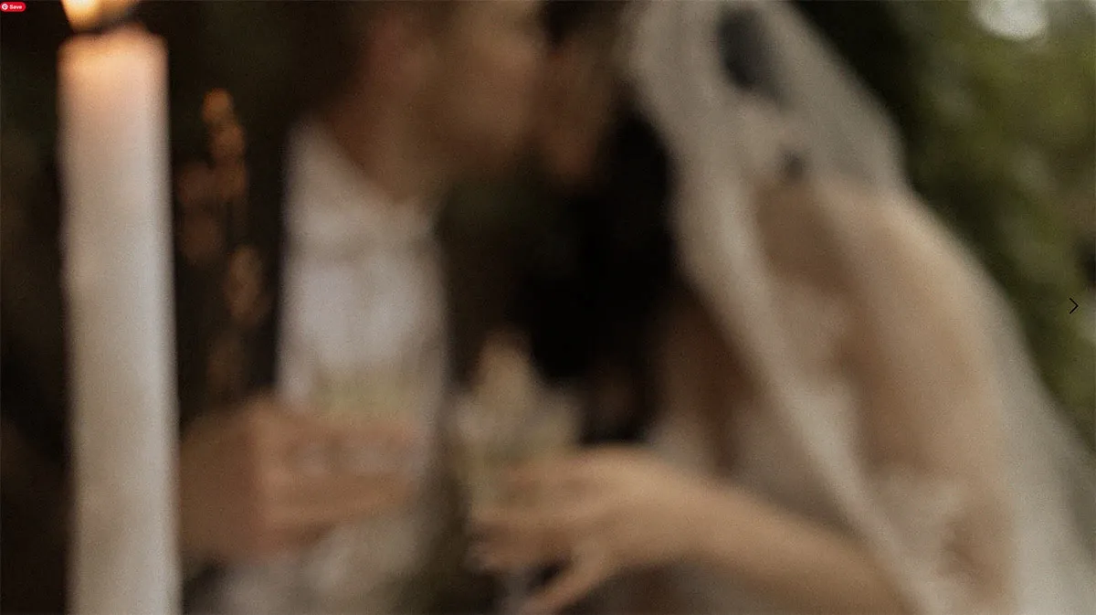 This image features a blurred view of a bride and groom sharing an intimate moment, creating an artistic and romantic effect. They are softly kissing, with the bride dressed in a flowing gown and veil, and the groom in a suit. Both are holding glasses, likely making a celebratory toast. A tall, elegant candle is situated nearby, adding to the wedding ambiance. The photograph captures a sense of intimacy and celebration, showcasing the wedding photography skills of Acrewood, a featured vendor on Mariage Elegant, Canada.