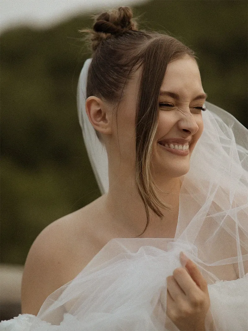 A smiling bride, with her hair styled in an elegant bun with a veil, is capturing a joyful moment outdoors, against a backdrop of lush greenery. She is dressed in a strapless wedding gown. This image showcases the wedding photography expertise of Acrewood, a vendor featured in the wedding planning and vendor listing at Mariage Elegant, Canada.