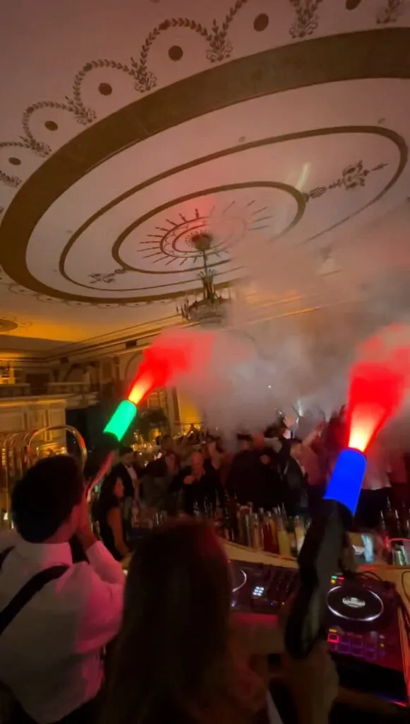 A lively wedding reception scene featuring PJ Entertainment, a wedding entertainment vendor, at Mariage Elegant, Canada. The image shows a DJ booth with a couple of DJs energetically entertaining guests. The DJs are using colorful LED smoke cannons, creating a vibrant and festive atmosphere. The elegant venue is adorned with an intricate ceiling design, contributing to the sophisticated ambiance. Guests are seen dancing and enjoying the celebration, highlighting the dynamic and engaging entertainment provided by PJ Entertainment.
