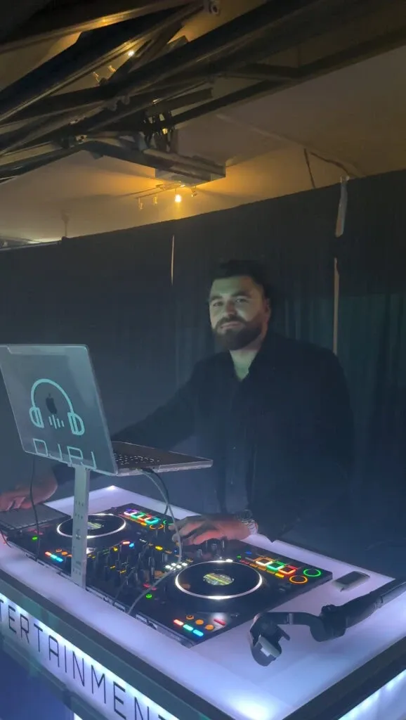 This image features a DJ from PJ Entertainment, a wedding entertainment vendor, performing at an event. The DJ is standing behind a lit DJ booth adorned with equipment such as a mixer and turntables. He is focused on the music, surrounded by ambient lighting that creates a vibrant atmosphere, perfect for wedding celebrations. The setting suggests a lively and engaging experience, highlighting PJ Entertainment's role in providing dynamic musical entertainment for weddings hosted by Mariage Elegant in Canada.