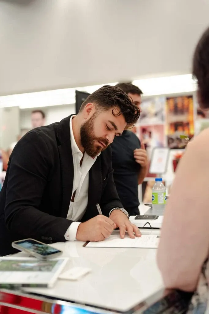 A professional DJ from PJ Entertainment, a wedding entertainment vendor, is seen attentively signing a contract at a vendor booth. The DJ, dressed in a black suit jacket and white shirt, is focused on the paperwork in front of him on a white counter. Nearby, a smartphone and a bottle of water are visible, emphasizing a modern and dynamic setting. In the background, blurred attendees and displays suggest a lively wedding expo environment, highlighting the importance of engaging wedding entertainment services.