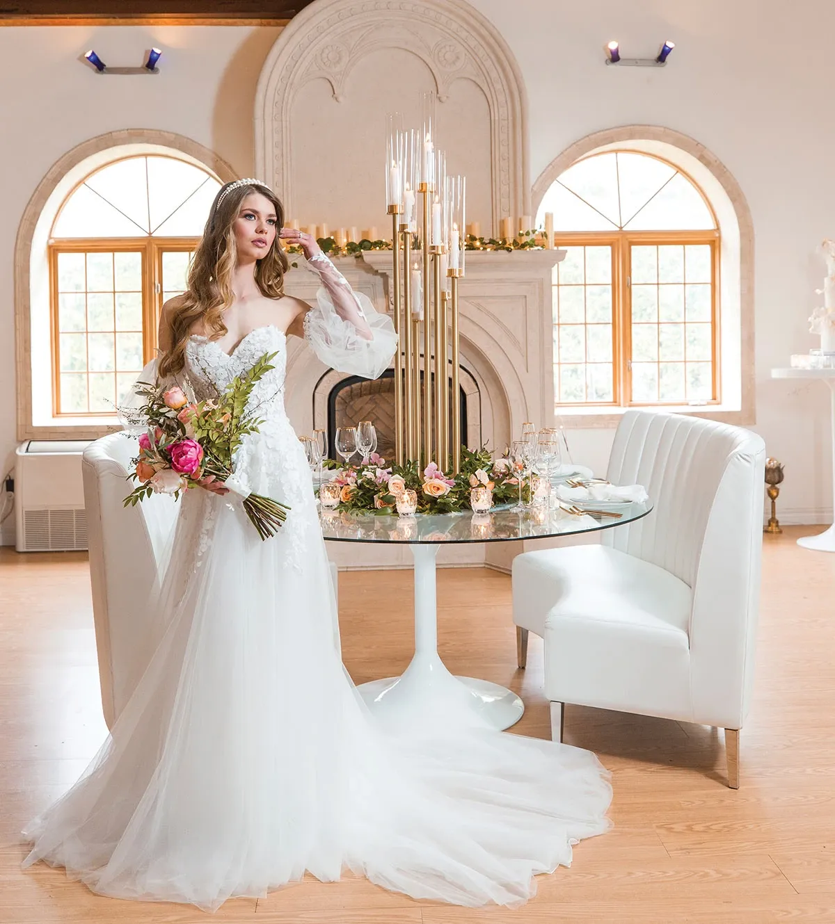 A bride in an elegant white wedding gown stands near a round table, holding a vibrant bouquet of pink and white flowers. The table is set elegantly with glassware and floral centerpieces, featuring soft pink roses. Behind her, a classic fireplace with tall, decorative candlesticks enhances the sophisticated ambiance of the room. The arched windows allow natural light to flood in, highlighting the luxurious setting. This image showcases Celebrations Group, a vendor in the Wedding Rentals category, demonstrating their ability to create a stylish and romantic wedding atmosphere.