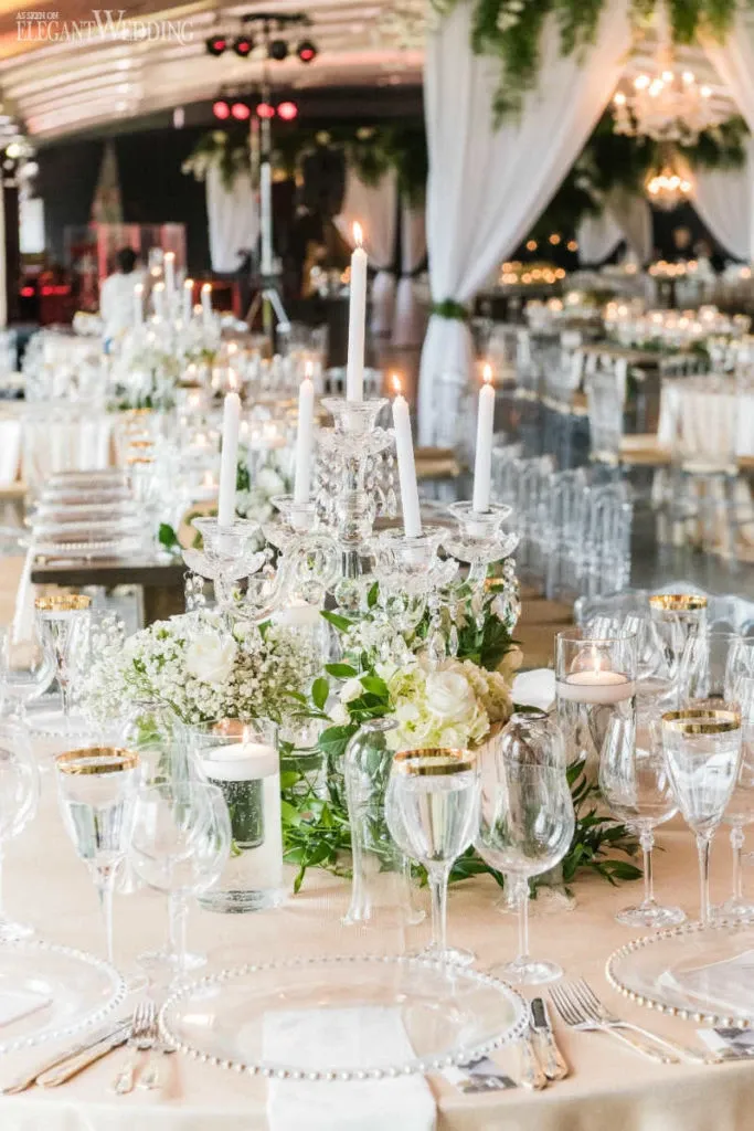 The image features a beautifully arranged wedding reception setup by Celebrations Group, a vendor in the Wedding Rentals category. The scene showcases an elegantly decorated table adorned with a stunning crystal candelabra at its center, surrounded by white flowers and lush greenery. Fine glassware, including wine glasses with gold rims, is pristinely arranged around the centerpiece. The table setting includes exquisite white plates with beaded edges and neatly folded napkins. In the background, draped white fabrics and chandeliers create a luxurious and romantic ambiance, perfect for a wedding celebration.