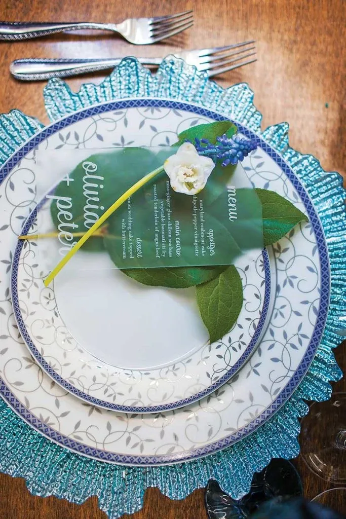 The image showcases an elegant table setting by Celebrations Group, a vendor in the Wedding Rentals category. The focus is on a beautifully arranged plate set featuring intricate blue and white floral patterns. A delicate, transparent menu rests on top, adorned with fresh greenery and a single white rose with a blue flower, adding a touch of natural elegance. Forks are neatly placed to the side, highlighting the sophisticated dining presentation perfect for a wedding occasion. The table is set on a wooden surface, enhancing the rustic and elegant theme suitable for any wedding event planned through Mariage Elegant in Canada.