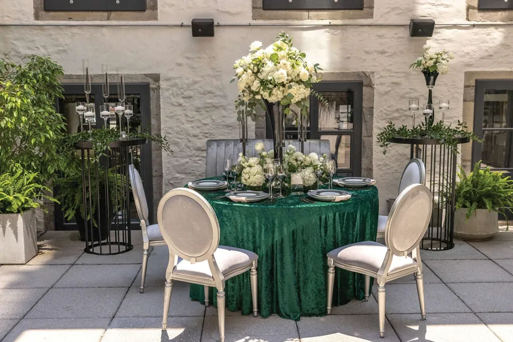 The image features a beautifully arranged outdoor wedding reception setup by Celebrations Group, categorized under Wedding Rentals. At the center of the scene is a round table adorned with a luxurious emerald green tablecloth. The table is elegantly set with fine china, cutlery, and glassware, surrounded by classic white upholstered chairs. A stunning floral centerpiece composed of white roses and greenery towers above the table, adding a touch of elegance. To enhance the ambiance, tall candle holders and additional floral arrangements are placed strategically around the area. The setting is located on a charming stone patio, surrounded by lush greenery, reflecting a sophisticated and romantic wedding atmosphere.