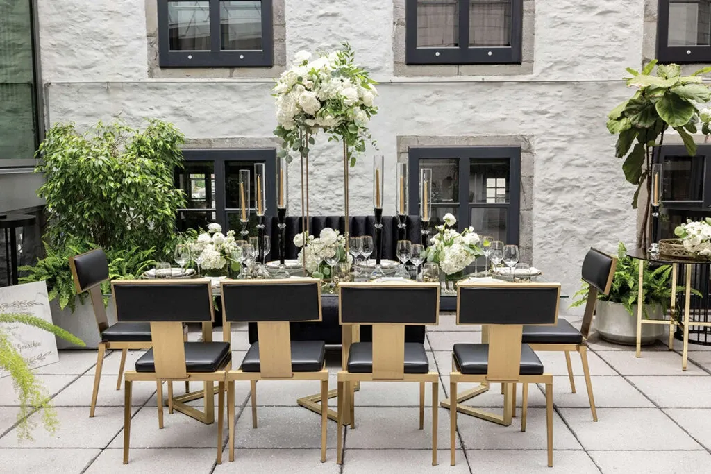 Elegant wedding reception setup by Celebrations Group, showcasing their wedding rentals category. The image features a beautifully arranged outdoor dining table set for an upscale wedding event. The table is adorned with sophisticated black-and-gold chairs and lavish floral centerpieces of white flowers and greenery. Tall, slender black candles add a touch of elegance, enhancing the sophisticated atmosphere. Set against a backdrop of a historic-style building with large windows and surrounded by lush plants, this arrangement exemplifies stylish and chic wedding decor.