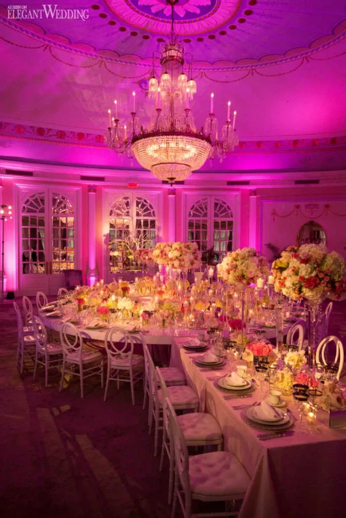 The image shows an opulently decorated wedding reception hall provided by Celebrations Group in the Wedding Rentals category. The venue features a large, elegant chandelier hanging from a domed ceiling, casting a warm glow over the space. The room is enhanced with soft pink and purple uplighting, creating a romantic ambiance. Long tables are adorned with white linens, gold-rimmed plates, and ornate glassware. Stunning floral centerpieces, featuring an array of white and pink flowers, add a touch of sophistication. White chairs with elegant designs surround the tables, completing the luxurious setup.