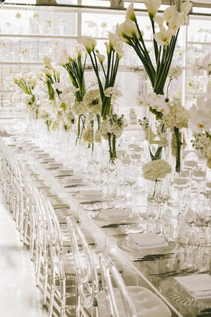 The image features a beautifully arranged wedding reception table set by Celebrations Group, a vendor specializing in wedding rentals listed on Mariage Elegant, Canada. The long, elegantly dressed table is adorned with tall, clear vases filled with white calla lilies and orchids, adding a sophisticated floral touch. Transparent chairs surround the table, enhancing the ethereal, modern aesthetic. Each place setting includes intricately folded white napkins and sparkling glassware, reflecting the natural light streaming into the airy venue. This setup exemplifies a stylish and graceful wedding ambiance.