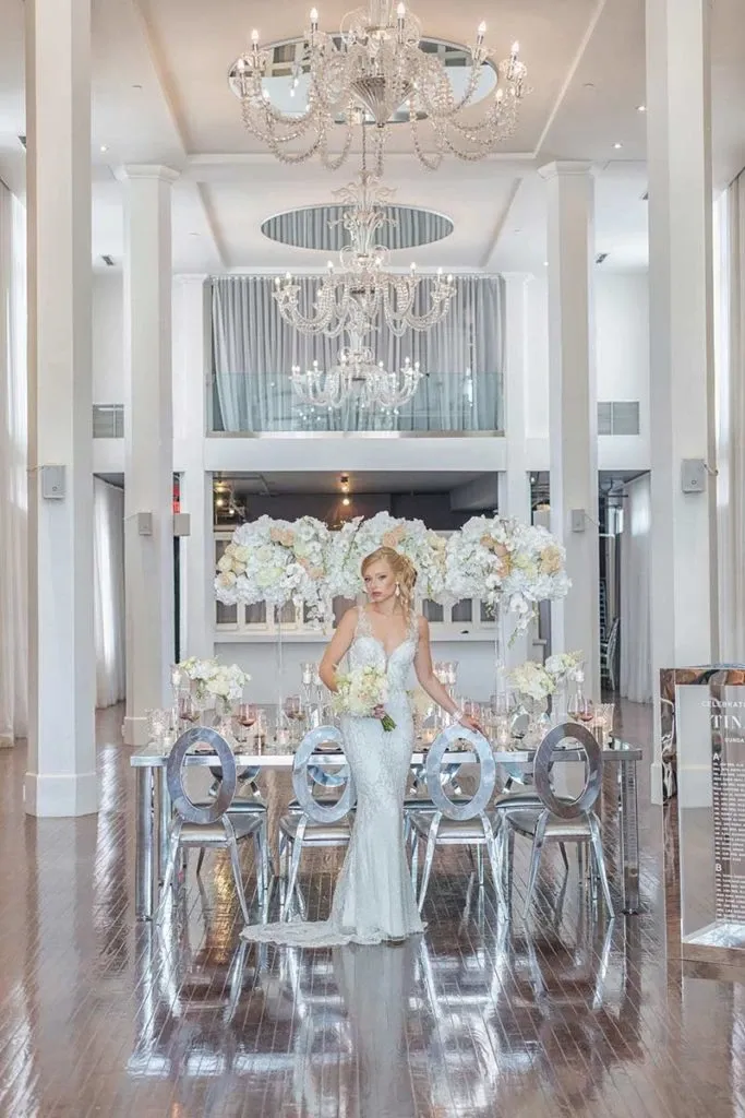 This image showcases an elegantly decorated wedding reception area provided by Celebrations Group, categorized under Wedding Rentals. The scene features a lavishly set table adorned with an array of white floral centerpieces, complemented by sleek, modern chairs. A bride dressed in a stunning white gown stands prominently in the center, adding a touch of elegance to the setting. The chandelier above casts a warm glow over the polished wooden floor and tall white columns, enhancing the luxurious atmosphere of the venue, ideally suited for a sophisticated wedding celebration in Canada.