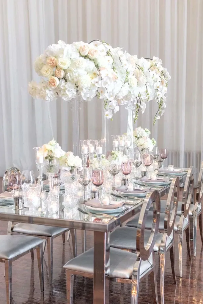 The image shows an elegant wedding reception table set up by Celebrations Group, featured in the wedding rentals category. The table is adorned with a stunning floral centerpiece, featuring a lavish arrangement of white and blush roses, along with cascading orchids and greenery. The mirrored table surface reflects the exquisite decor, and the setting includes silver-rimmed glassware, crystal candle holders, and neatly folded napkins. The chairs have a sleek metallic finish, enhancing the sophisticated ambiance, perfect for a luxurious wedding event by Mariage Elegant in Canada.
