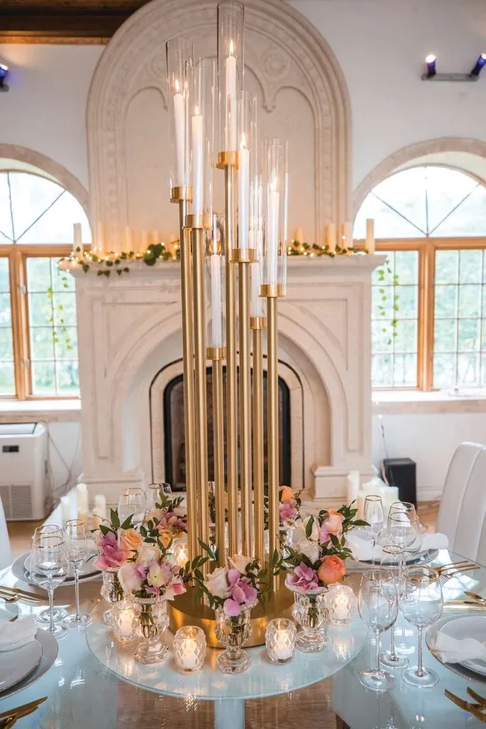 The image features an elegantly set table by Celebrations Group, categorized under Wedding Rentals, at a beautifully decorated venue. A tall, gold candle holder with multiple slender candles serves as the centerpiece, surrounded by a lush arrangement of pastel flowers, including pink and peach blooms. The table is set with fine glassware and plates, enhancing the sophisticated aesthetic. In the background, there is a grand fireplace adorned with greenery and candles, adding a romantic ambiance, alongside large windows allowing natural light to fill the space, creating a perfect setting for a wedding celebration.