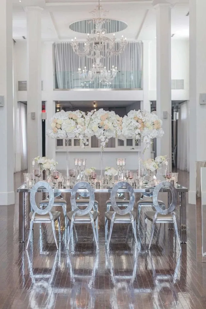 The image features an elegant wedding setup by Celebrations Group, categorized under Wedding Rentals. It showcases a lavish dining table adorned with tall, exquisite floral arrangements composed of white and blush flowers. The table is set with polished silver chairs, adding a modern and sophisticated touch. Candles in various sizes are symmetrically placed, enhancing the romantic ambiance. The setting is in a spacious, light-filled room with glossy wooden floors and a grand chandelier overhead, reflecting luxury and elegance typical of a wedding venue in Canada.