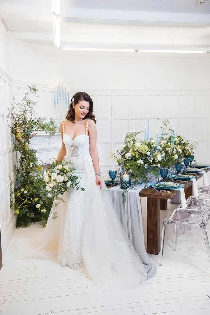 A bride in a white lace wedding gown stands beside an elegantly arranged reception table in a well-lit room. The table, provided by Celebrations Group under the Wedding Rentals category, is adorned with tall blue candles, lush green and white floral arrangements, and intricate tableware, including blue glass goblets and decorative plates. Clear ghost chairs surround the table, adding a modern touch to the classic setup. The backdrop features white-paneled walls and a decorative fireplace draped with greenery, creating a sophisticated and romantic atmosphere suitable for a wedding.