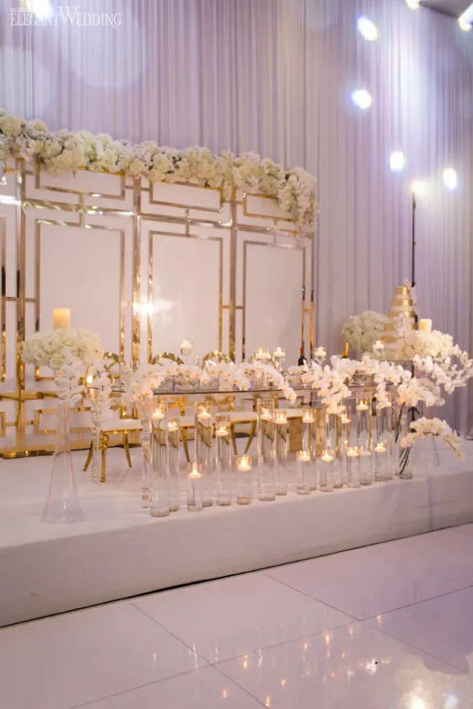 This image showcases a sophisticated wedding reception setup by Celebrations Group, a vendor in the Wedding Rentals category at Mariage Elegant, Canada. The scene features an elegant arrangement with an opulent white and gold theme. The backdrop consists of beautifully reflective panels framed in gold, accented by lush, cascading white floral arrangements on top. In the foreground, a selection of tall, clear glass vases contains white orchids, while smaller vases house glowing candles, creating a romantic ambiance. A tiered wedding cake adorned with intricate white decorations is visible to the right, enhancing the luxurious setting. The scene is bathed in soft lighting, adding to the overall elegance of the display.