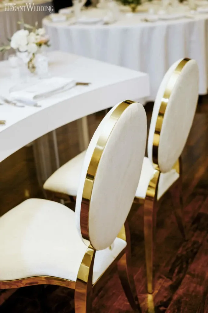 The image showcases an elegant reception setup provided by Celebrations Group, a vendor in the Wedding Rentals category. It features two luxurious white chairs with gold trim, positioned beside a white, curved table set with white napkins and ivory floral arrangements. The polished wood floor complements the sophisticated and modern aesthetic, ideal for a refined wedding setting in Canada.