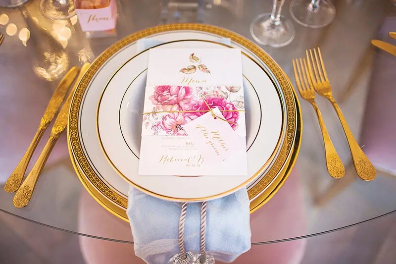 This image showcases an elegantly arranged table setting featuring exquisite wedding rentals from Celebrations Group, under the Wedding Rentals category. A luxurious gold-rimmed plate is set atop a glass table, accompanied by intricately designed gold cutlery. The centerpiece of the setting is a beautifully printed floral menu card and a tag, both resting on a folded light blue cloth napkin. Crystal glasses are placed behind the setting, contributing to the sophisticated and romantic ambiance appropriate for an upscale wedding event.