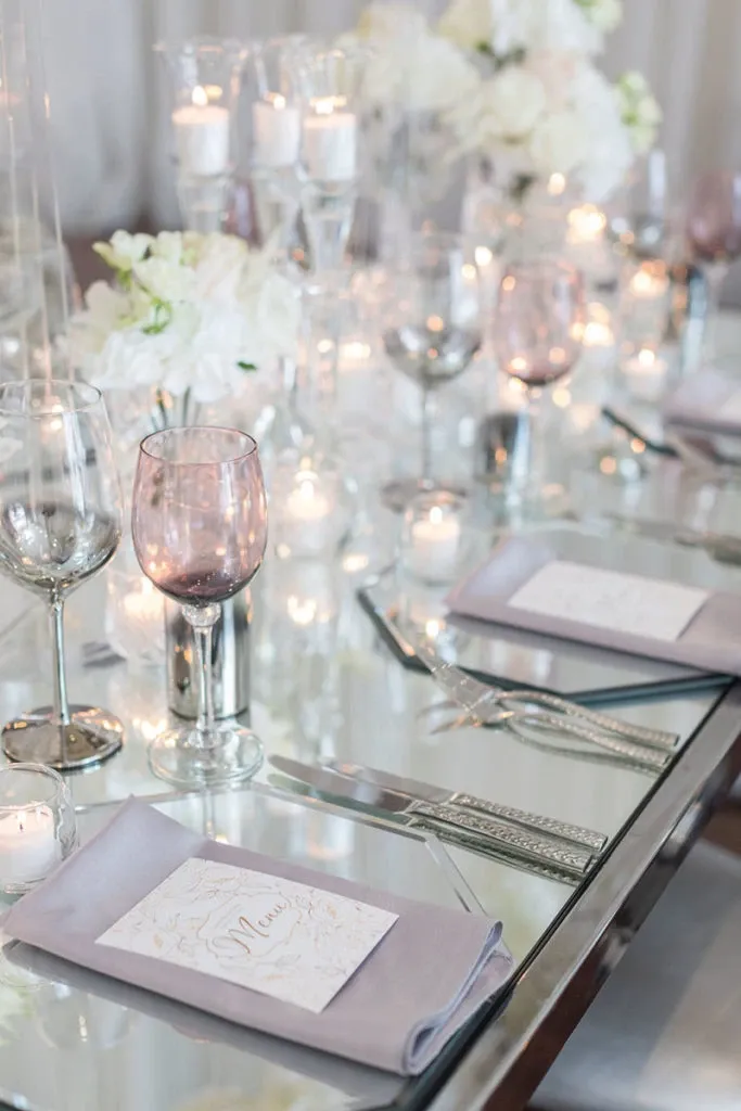 The image shows an elegantly set table provided by Celebrations Group, a vendor specializing in wedding rentals. The table features a mirrored surface reflecting soft candlelight, enhancing the romantic ambiance. Delicate lavender napkins hold beautifully designed menu cards, while silver cutlery and wine glasses are neatly arranged. Crystal candle holders and vases with white floral arrangements add sophistication, making it an ideal setup for a luxurious wedding reception.