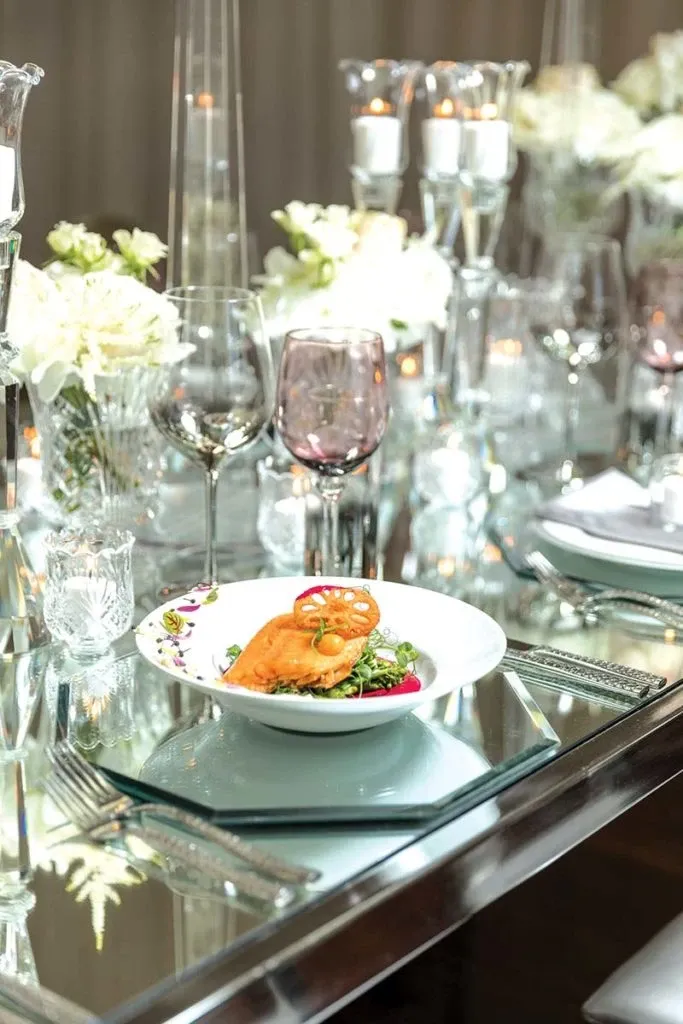 The image showcases an elegant wedding reception table set up by Celebrations Group, a vendor specializing in Wedding Rentals. The table features a reflective glass surface adorned with intricate floral arrangements of white roses and greenery. Crystal candle holders with lit candles add a romantic ambiance. Each place setting includes elegantly arranged glassware, including wine glasses with a subtle tint. A stylishly plated dish with gourmet food sits on a mirrored charger, adding a touch of sophistication to the overall display.