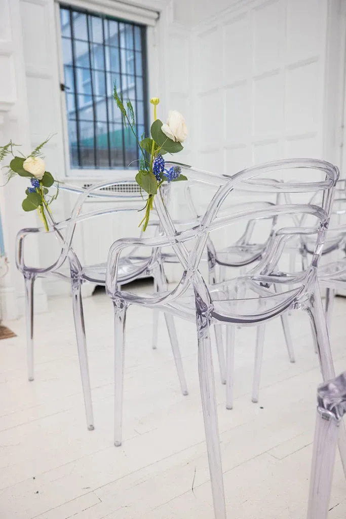 Transparent, modern acrylic chairs from Celebrations Group are neatly arranged in a bright, white interior space. Each chair features elegant floral decorations with white and blue flowers and greenery, offering a chic and sophisticated setup for a wedding ceremony. The large window in the background allows natural light to fill the room, highlighting the stylish and minimalistic design. Perfect for couples seeking contemporary wedding rentals from Celebrations Group, part of Mariage Elegant in Canada.
