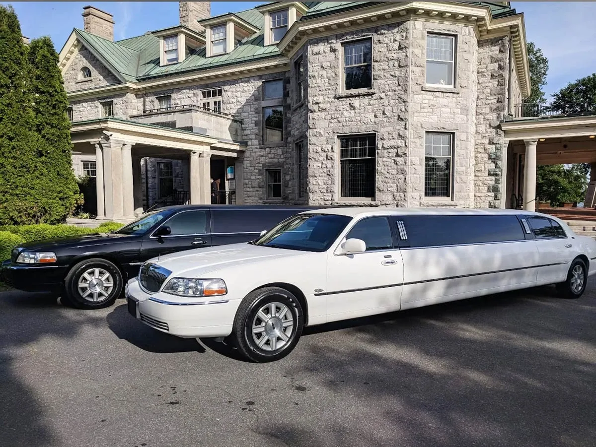 This image features two luxurious limousines from Ambiance Limousines, under the wedding category of Wedding Limousine, parked elegantly in front of a grand stone mansion, perfect for a wedding backdrop. The limousines, one black and one white, are sleek and polished, ideally suited for wedding transportation. Lush greenery and a clear blue sky complement the elegant setting, enhancing the ambiance for a sophisticated wedding arrival or departure.