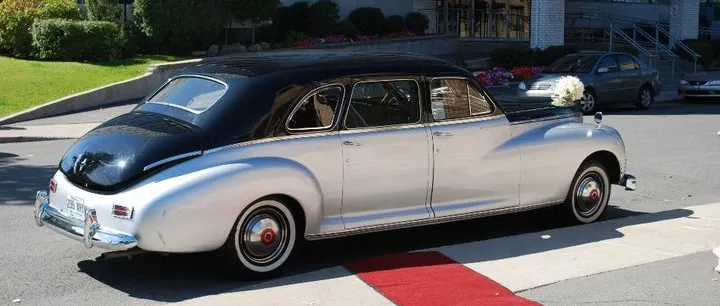In the image, a classic vintage limousine provided by Ambiance Limousines, under the wedding limousine category, is elegantly parked on a vibrant red carpet. The car features a sleek, two-tone black and silver exterior with gleaming chrome details, embodying a sense of luxury and sophistication suitable for a wedding. A charming white floral arrangement adorns the side mirror, adding a romantic touch to the vehicle's presentation. The limousine is parked in an outdoor area, with a grassy backdrop and parked cars visible, complementing its elegant appearance for a memorable wedding entrance or exit.