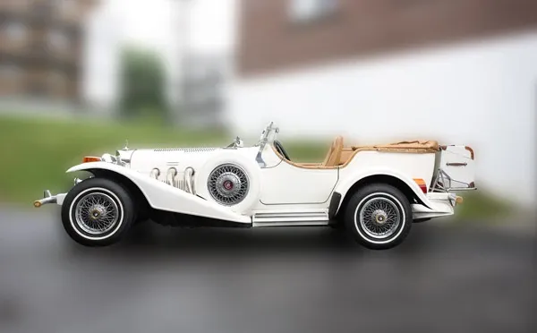 The image showcases a vintage-style wedding limousine from Ambiance Limousines, categorized under Wedding Limousine services. This elegant vehicle features a sleek white exterior with intricate chrome detailing, wire-spoke wheels, and a convertible leather top. The classic design adds a timeless charm, perfect for a sophisticated wedding entrance. The background is slightly blurred, highlighting the limousine as the central focus, ideal for capturing attention on the Mariage Elegant vendor listing in Canada.