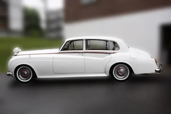 A classic white limousine provided by Ambiance Limousines, showcased for the wedding category on Mariage Elegant, Canada. The vintage vehicle is elegantly parked outdoors, featuring sleek lines and a polished surface, perfect for sophisticated wedding transportation. A subtle floral decoration adorns the front of the limo, adding a touch of wedding charm to its timeless design. The background is softly blurred, keeping the focus on the luxurious limousine.
