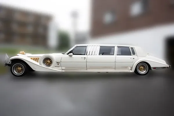 The image features an elegant, vintage-style white limousine provided by Ambiance Limousines, categorized under Wedding Limousine services. The limousine showcases intricate gold detailing on the front and a sleek, elongated body design, perfect for a luxurious arrival at wedding events. It is parked outdoors against a blurred background, emphasizing its classic and stylish appearance. This vehicle adds a touch of sophistication and grandeur to any wedding celebration, aligning with the offerings of Mariage Elegant in Canada.
