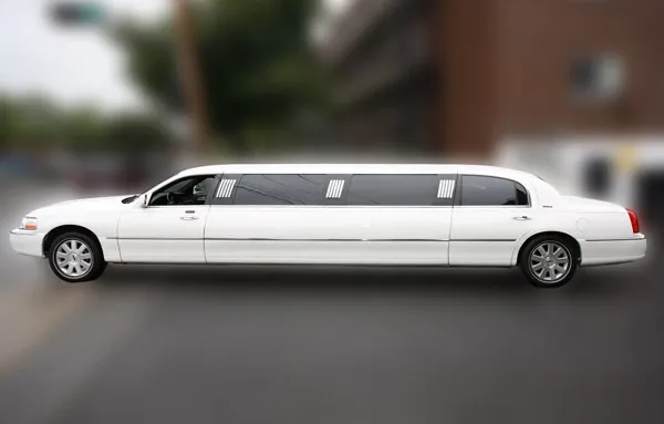 The image shows an elegant, white stretch limousine provided by Ambiance Limousines, featured in the Wedding Limousine category on Mariage Elegant, Canada. The limousine is parked on a street and is sleek and well-polished, showcasing its luxury and spaciousness, ideal for wedding transportation. The background is slightly blurred to emphasize the vehicle, highlighting its clean design and intricate detailing suitable for a sophisticated wedding experience.