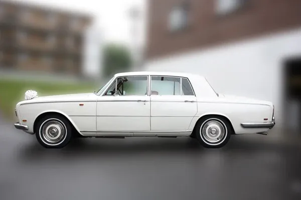 The image showcases a classic white limousine from Ambiance Limousines, a vendor listed under the Wedding Limousine category on Mariage Elegant, Canada. The elegant vehicle is parked against a blurred urban background, emphasizing its sleek, vintage design. The limousine is polished to a high shine, with chrome accents and smooth lines, making it an ideal choice for a sophisticated wedding entrance. The timeless style and pristine condition of the limousine suggest a commitment to luxury and quality, perfect for transporting the bridal party in style.