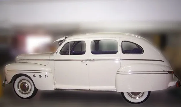 A vintage, white limousine is showcased, featuring a smooth, elegant design typical of classic cars, ideal for a wedding setting. The vehicle, provided by Ambiance Limousines, represents the wedding limousine category. It has polished chrome accents and whitewall tires, adding to its timeless sophistication. The windows are tinted for privacy, making it a fitting choice for a stylish and memorable entrance or exit for wedding couples.