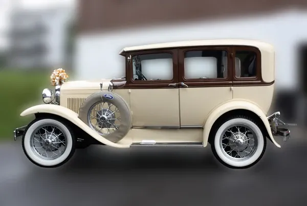 The image features a vintage ivory and brown limousine by Ambiance Limousines, categorized under Wedding Limousine, suitable for wedding events. The classic vehicle is adorned with elegant floral decorations on the front, showcasing a luxurious and timeless style perfect for sophisticated weddings. The intricate wheel design and pristine condition of the car highlight its elegance, adding a touch of glamour to any wedding celebration. The blurred background emphasizes the focus on the limousine, making it an attractive option for couples seeking a unique and stylish arrival on their special day.