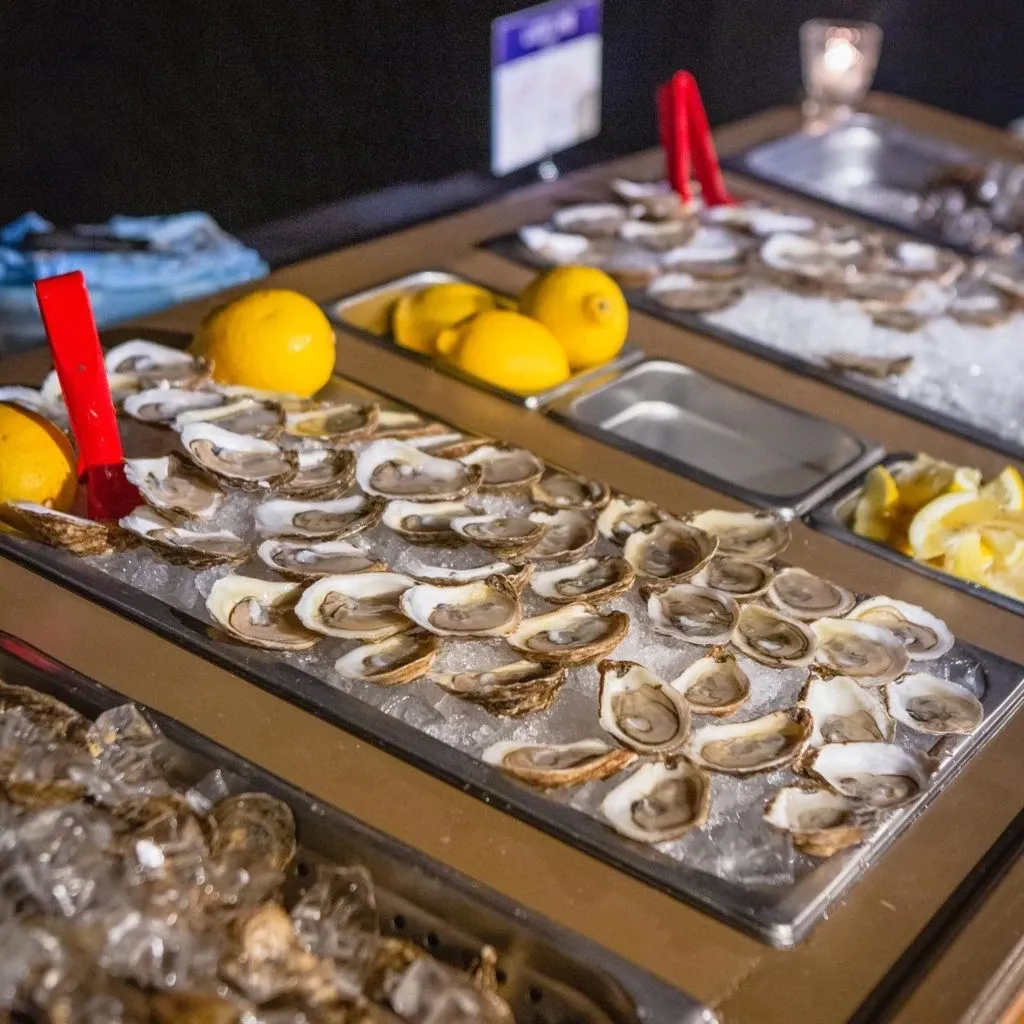 This image shows an elegantly arranged oyster bar by Oysters In Motion, categorized under Wedding Catering on Mariage Elegant, Canada. The display features a variety of fresh oysters placed on a bed of ice, garnished with vibrant lemon wedges for added flavor. Two red serving tongs are visibly positioned for easy access to the oysters. The setup is completed with a softly lit candle in the background, enhancing the sophisticated ambiance suitable for a wedding event.