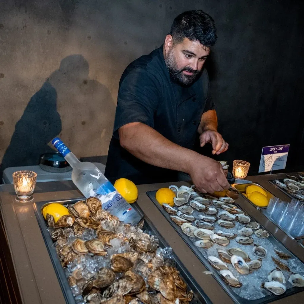 In the image, a chef from Oysters In Motion, a wedding catering vendor, is skillfully preparing fresh oysters at a wedding event hosted by Mariage Elegant in Canada. He is standing behind a well-organized display featuring trays filled with ice and an assortment of shucked oysters, ready for serving. Sliced lemons are strategically placed among the oysters for added flavor. A bottle of Grey Goose vodka is visible, suggesting an elegant touch to the catering setup. The dimly lit setting is enhanced by small candles, creating a warm and inviting atmosphere, appealing for a festive wedding reception.