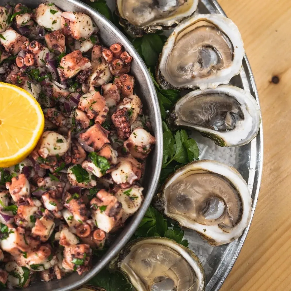 A beautifully arranged platter from Oysters In Motion, a wedding catering vendor, featuring fresh oysters on half shells encircling a stainless steel dish filled with a vibrant octopus salad. The salad is garnished with fresh herbs and a lemon half, providing a colorful contrast to the seafood. This elegant presentation is perfect for a sophisticated wedding reception, showcasing the vendor’s dedication to quality and aesthetics.