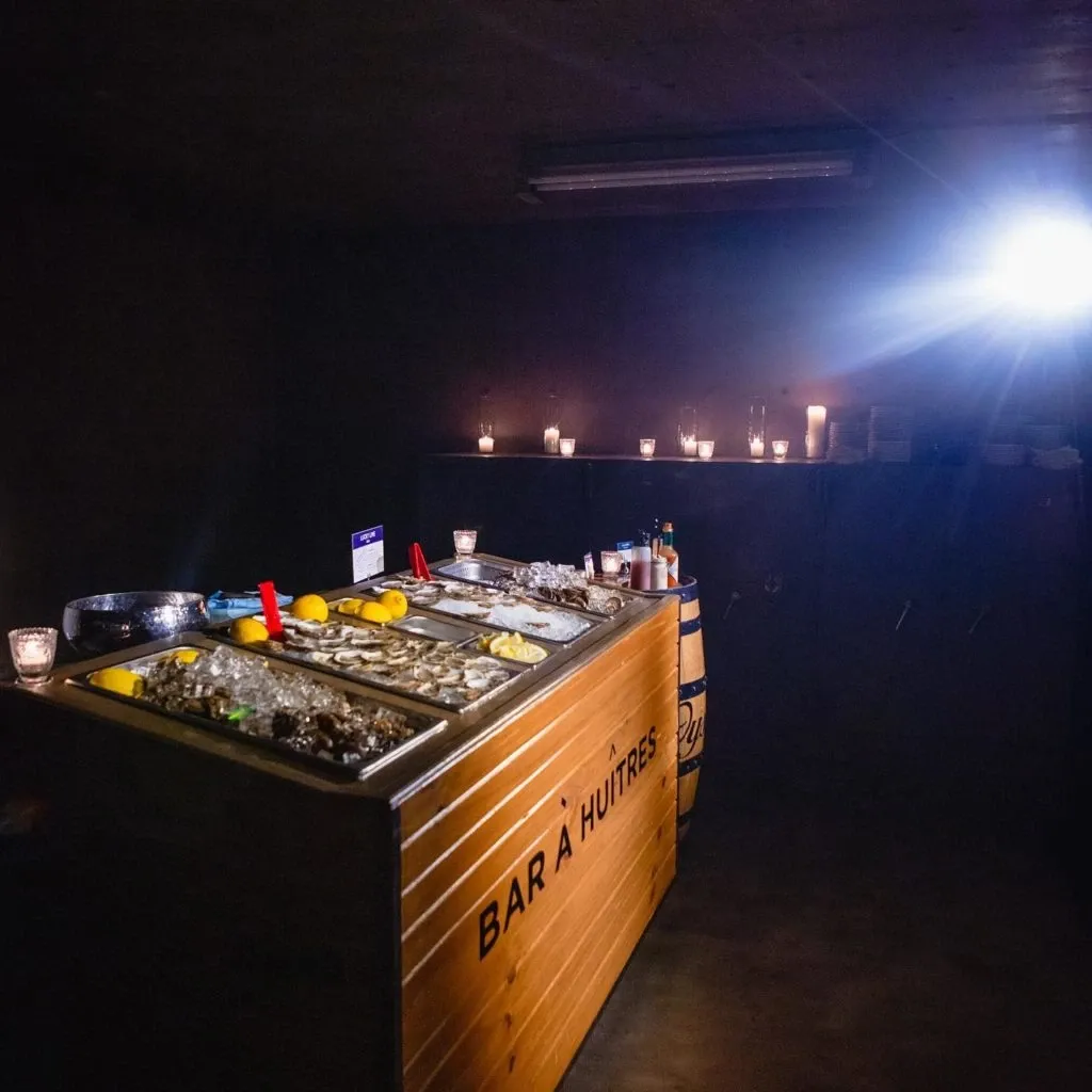 The image shows an elegant oyster bar setup by Oysters In Motion, categorized under Wedding Catering, for the Mariage Elegant vendor listing in Canada. The bar, labeled 'Bar à Huîtres,' is made of polished wood and displays a variety of fresh oysters on ice, accompanied by lemon wedges and sauces. The ambiance is set by softly lit candles placed along the back wall, creating a warm and inviting atmosphere suitable for wedding receptions. This setup highlights the sophisticated and professional presentation offered by Oysters In Motion for special events.