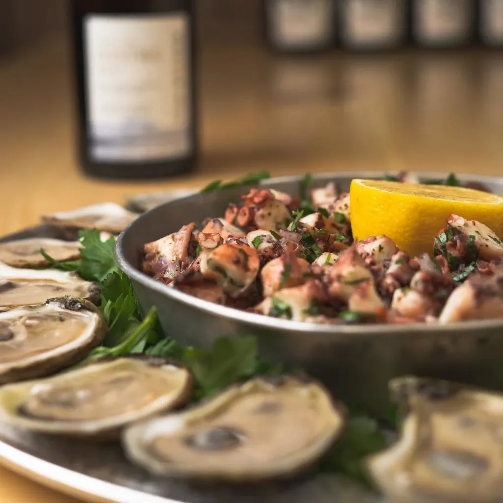 A beautifully arranged platter from Oysters In Motion, a wedding catering vendor at Mariage Elegant, Canada, featuring freshly shucked oysters displayed on a large metal tray. The oysters are garnished with sprigs of fresh parsley and accompanied by a central bowl of marinated seafood salad, topped with a lemon wedge. In the background, a blurred bottle of wine adds an elegant touch, suggesting a sophisticated dining experience perfect for a wedding celebration.