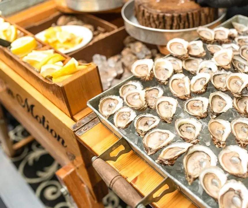 The image shows a beautifully arranged catering setup by Oysters In Motion, a wedding catering vendor. It features a wooden cart displaying a large tray of fresh oysters on a bed of ice, ready to be served. To the side, there are compartments containing lemon wedges and a variety of garnishes, enhancing the presentation. The elegant setup suggests a sophisticated and fresh addition to a wedding menu, highlighting the vendor's specialty in providing seafood delicacies for wedding events.