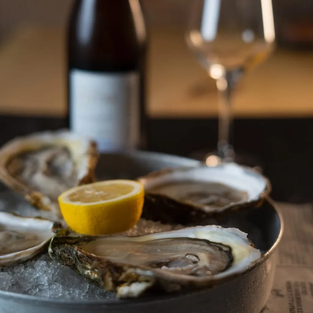 This image features a beautifully arranged display by Oysters In Motion, a wedding catering vendor. The presentation includes three half-shell oysters nestled on a bed of crushed ice in a round dish. A lemon wedge is placed prominently in the center, adding a touch of color and freshness. In the background, there is a blurred image of a wine bottle and an elegant wine glass, suggesting a refined dining experience suitable for weddings. The overall setting conveys a sense of sophistication and gourmet quality, ideal for upscale wedding events in Canada.