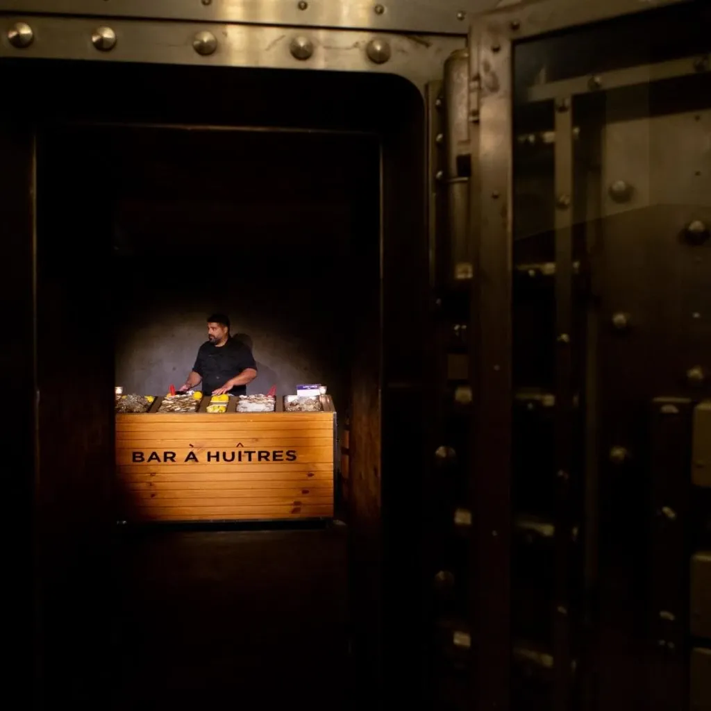 A chef from Oysters In Motion is standing behind a wooden seafood bar labeled 'Bar à Huîtres' inside a dimly lit room resembling a vault, showcasing their wedding catering service. The display features an assortment of fresh oysters and lemons, highlighting the vendor's specialty in providing elegant and upscale catering for weddings in Canada. This image captures the essence of sophisticated and unique culinary offerings available for wedding receptions.